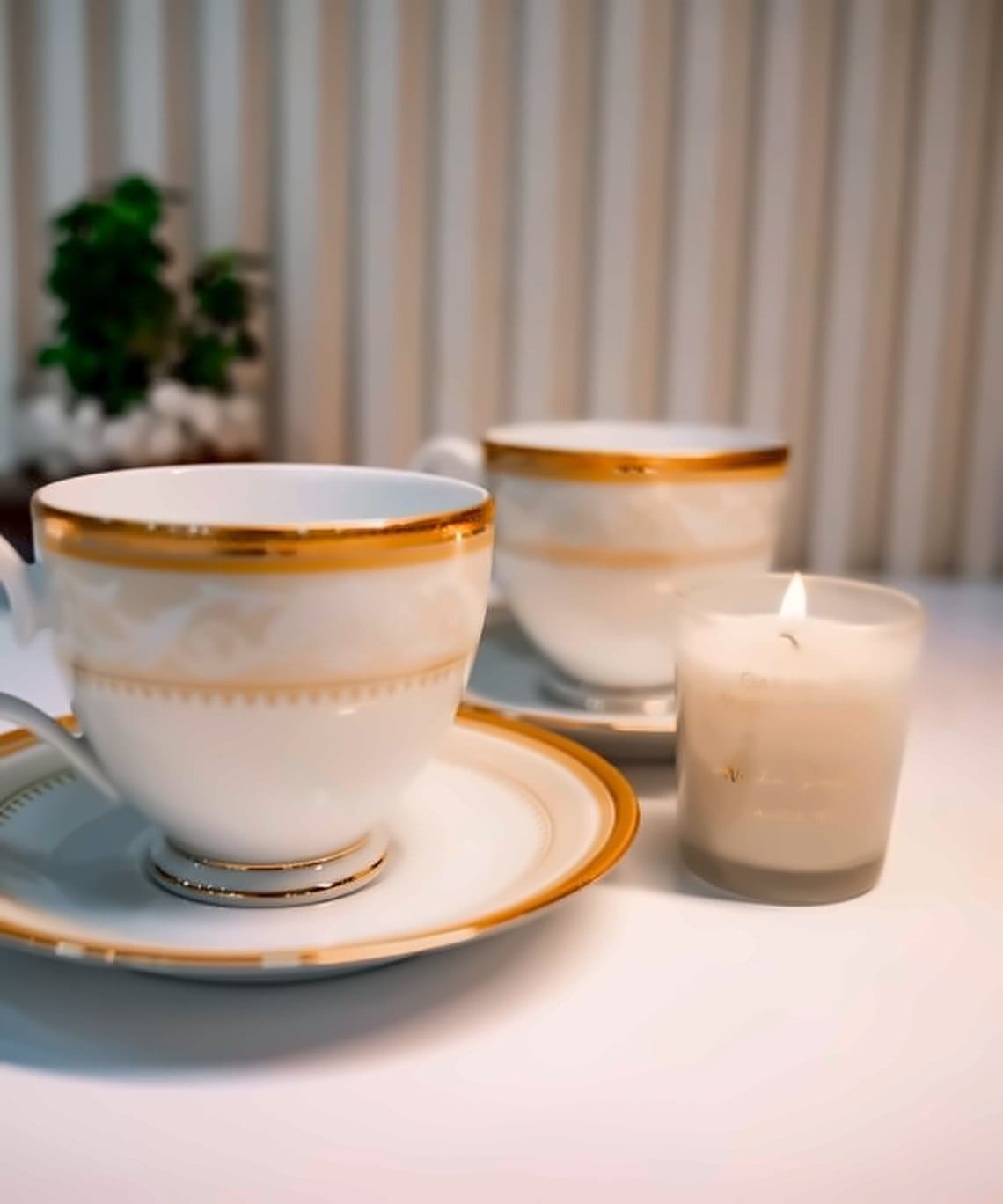 Two bone china tea cups with a gold baroque band on matching saucers beside a candle